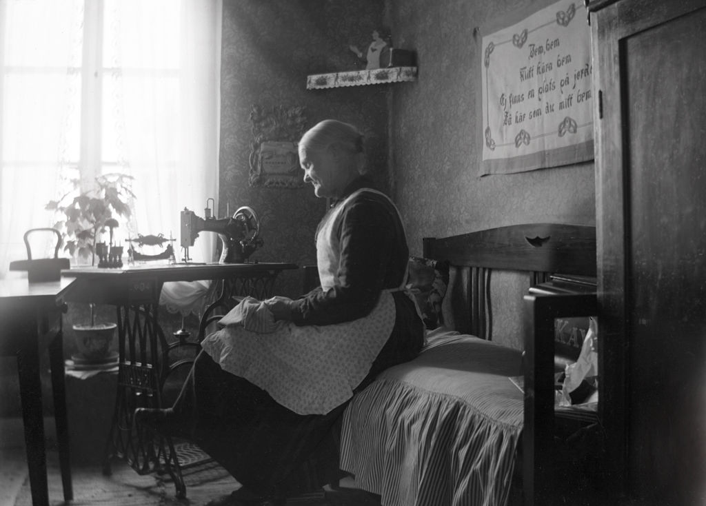 The woman at the sewing machine is Emelie Castensson (born 1854), who married with a slipper-maker Johan Andersson. The couple lived here when the picture was taken in Vagnsvik, Frinnaryd.