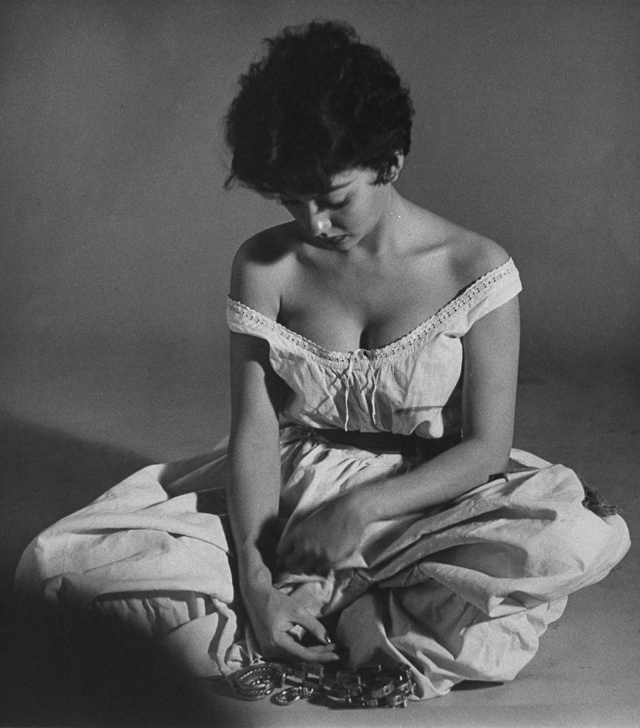 Rita Moreno sitting in an Indian squat looking coyly over her bare shoulder in front of a few trinkets on the ground as she plays girl rejected by her lover, 1954.