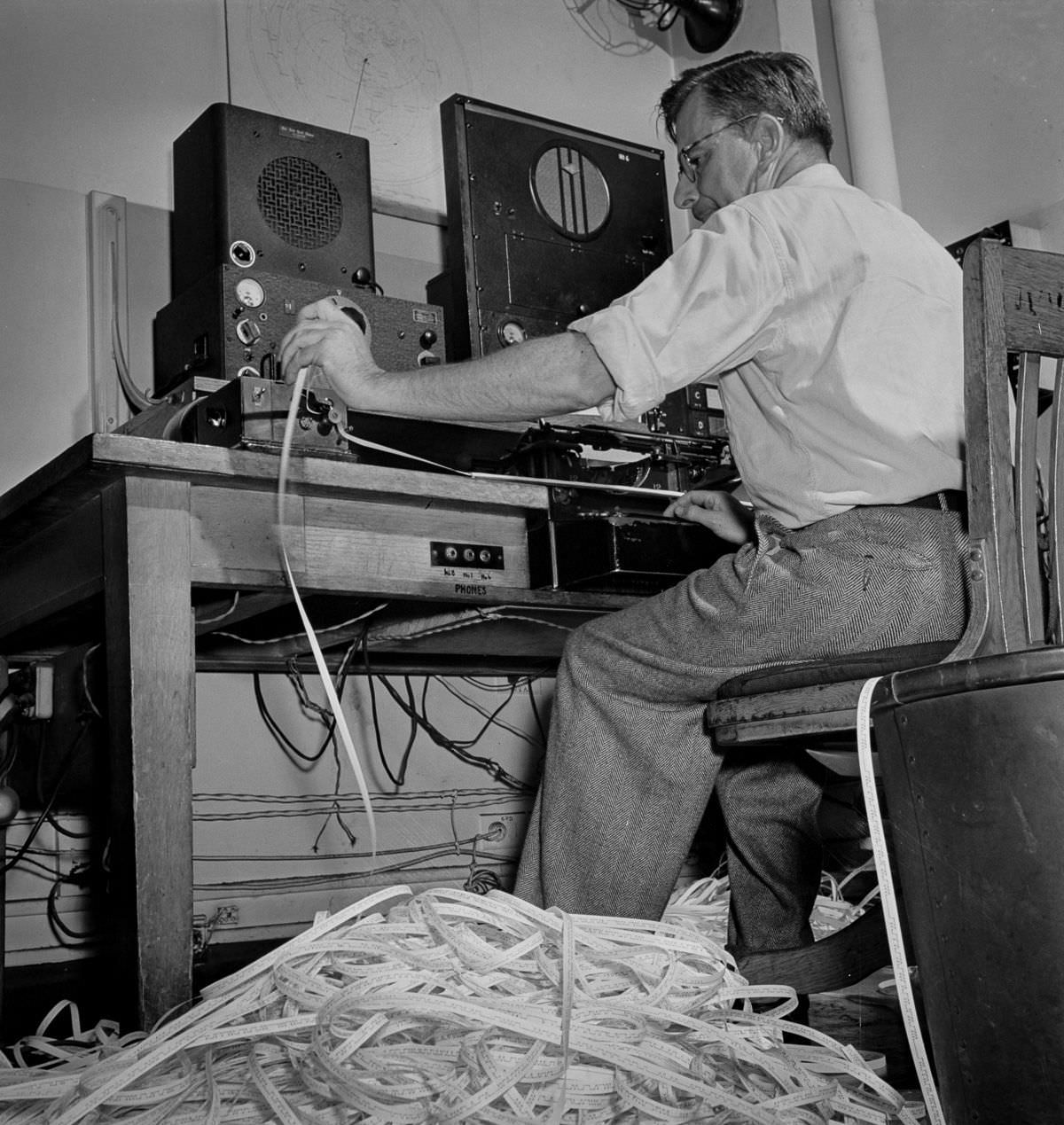 A radio operator listens in to Axis propaganda broadcasts. The paper piled on the floor has been examined to see what has already been covered in the last edition.