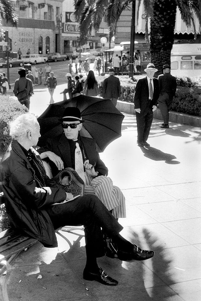 Two men sitting on park bench sheltering from the sun under an umbrella, San Francisco California, 1970.
