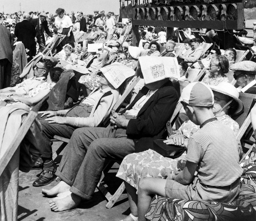 Two gentleman sitting on a busy Blackpool promenade in Lancashire, wearing newspapers on their heads as improvised sun hats, 26th July 1952.