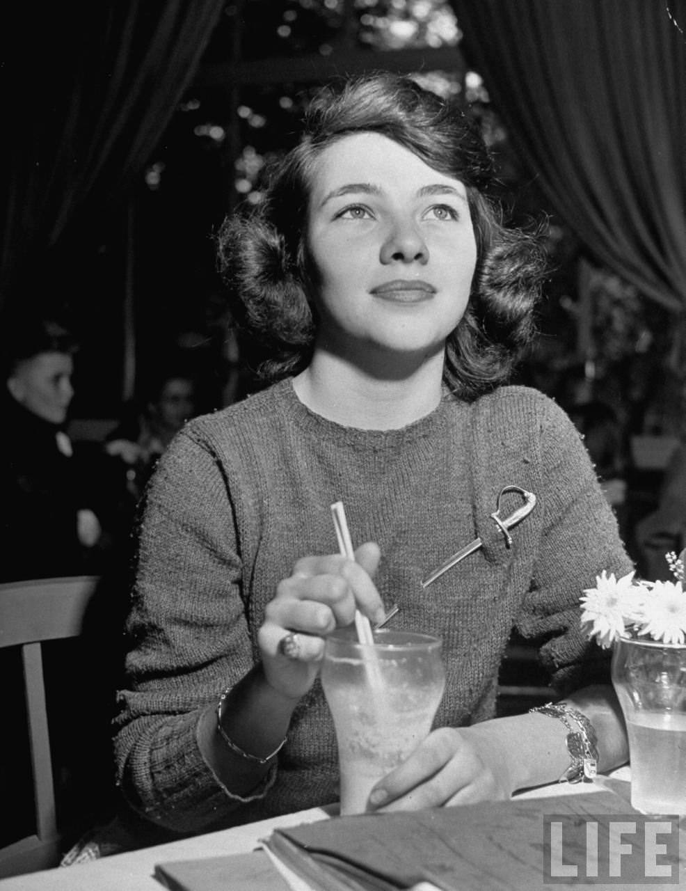 Margaret Raymond, 17, makes with a milkshake and models the fad of wearing miniature Heidelberg saberson sweater.