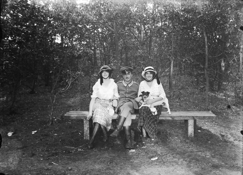 Bench in the woods, with a German soldier flanked by two women. The woman on the right has a dog on her lap, 1916