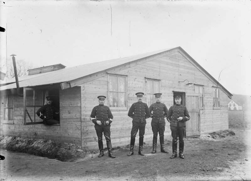 Four NCOs standing in front of a barracks. A fifth NCO sits in a window of the barracks (left).