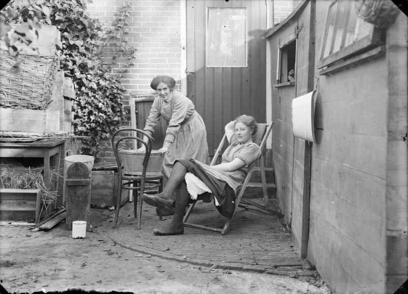 Two women in a place behind a house. One of them is doing the laundry in a zinc tub, the other is lying in a deck chair, 1916