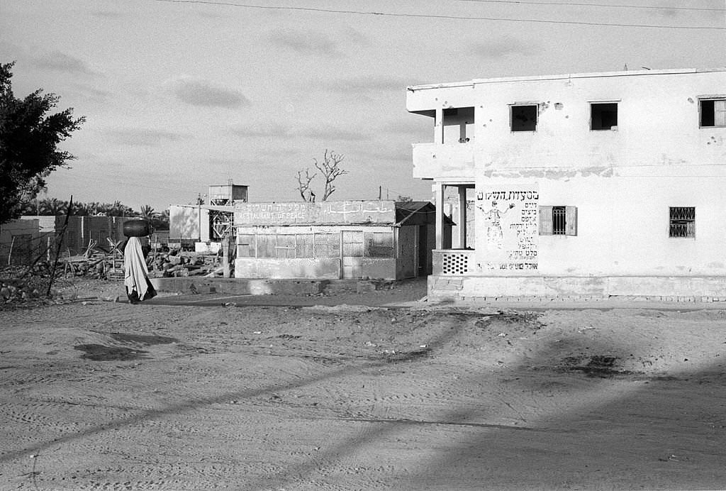 An Egyptian woman transports a water cistern on her head in a desolated background of the war passage, 1973