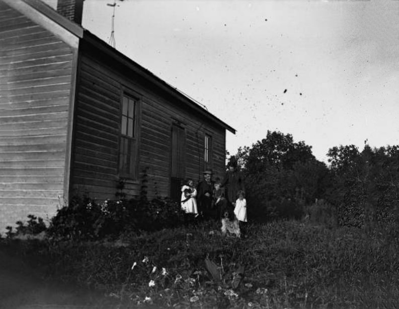 Family poses in front of frame house, 1900