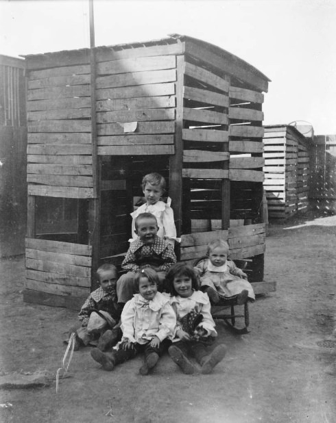 Childrens sit for camera beside chicken coop, 1900