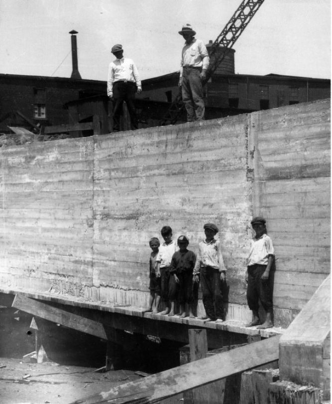 Five boys standing along the base of a poured concrete wall at a construction site, 1900