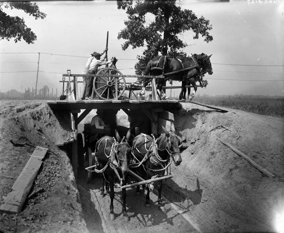 A couple of two-hitch horse wagons. The contents of one wagon is being poured into the wagon under the bridge, 1900
