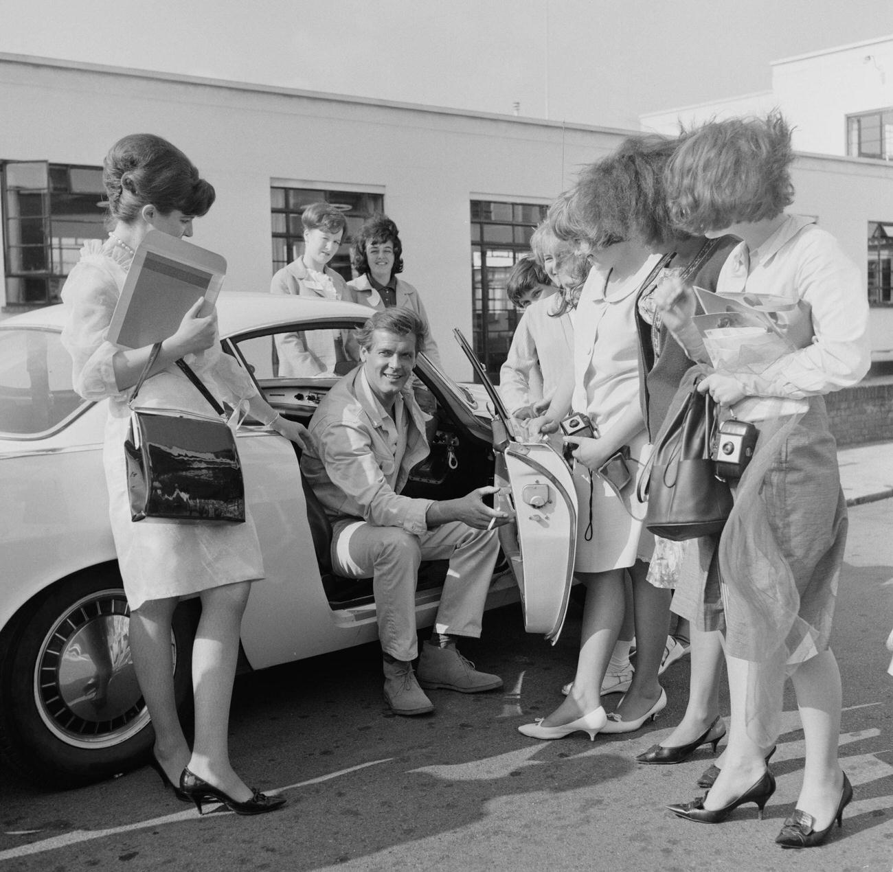 Roger Moore posed with female fans whilst seated in his Volvo P1800 car at Elstree Studios in Hertfordshire in August 1964.