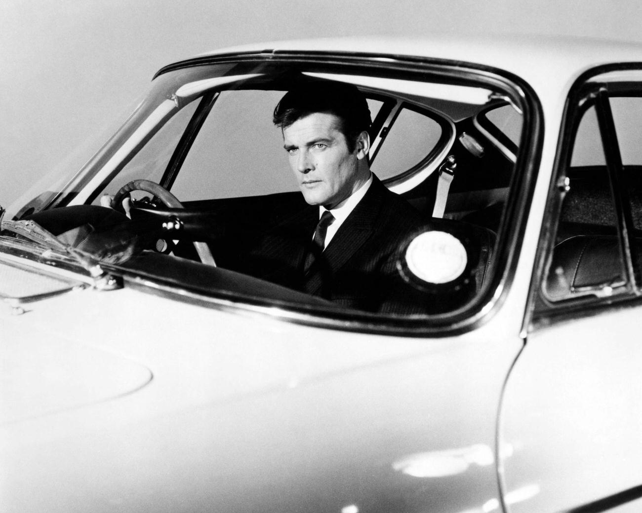 Roger Moore in a promotional portrait at the wheel of a Volvo P1800 sports car, for the British spy thriller TV series 'The Saint', 1965