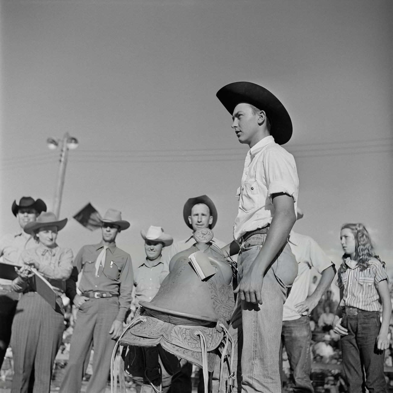A competitor holding his saddle at a youth rodeo in Houston, Texas, with other competitors and judges in the background, 1952.