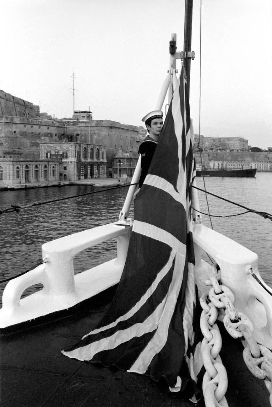 Radio operator Brian Houston covers the Union Jack at sunset in H.M.S. Blake in Valletta, Malta, 1972.