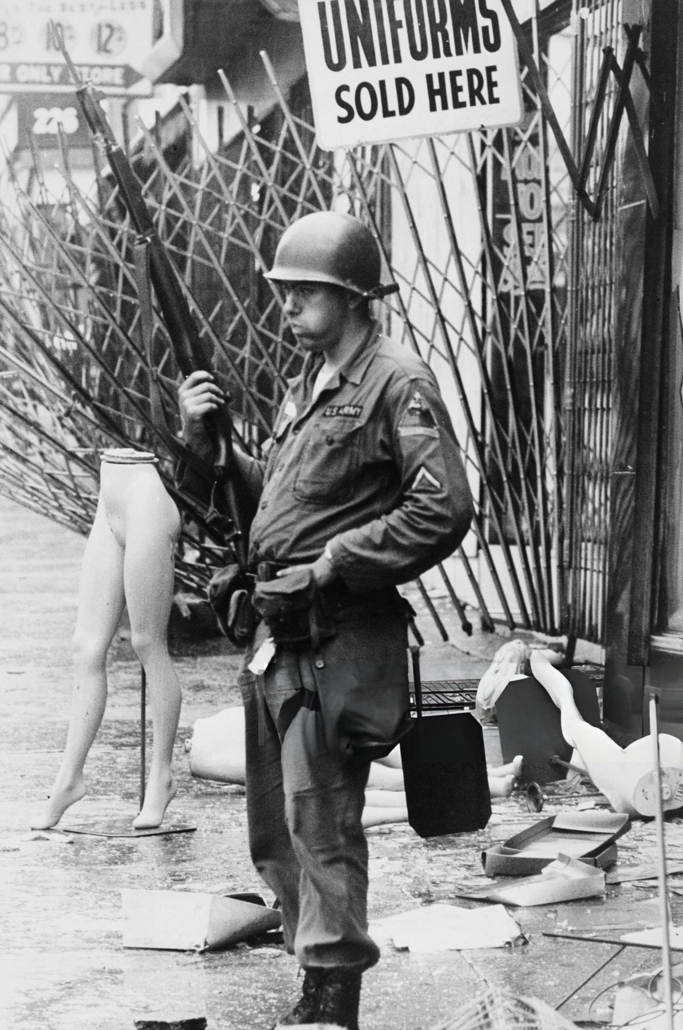 A soldier of the National Guard stands next to a mannequin's legs and a sign reading 'Uniforms Sold Here'