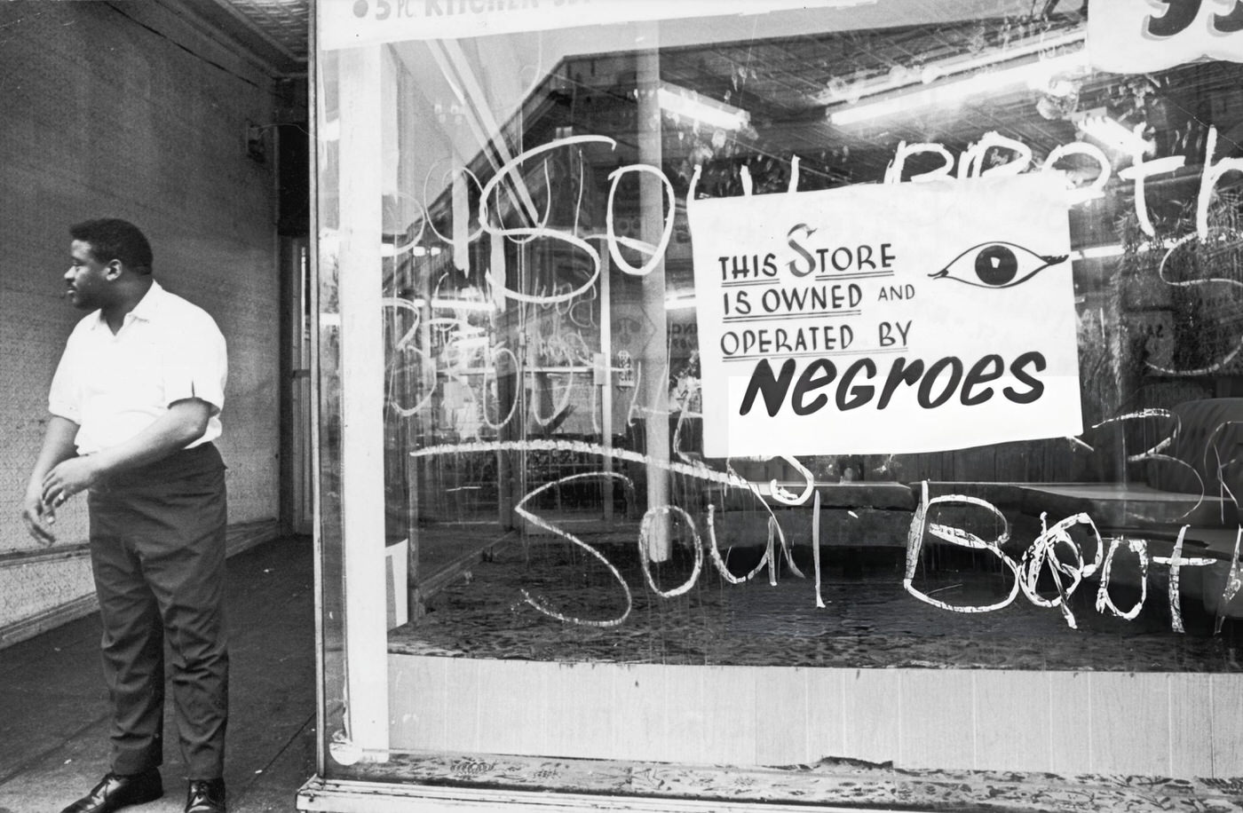 An African-American storekeeper stands outside his shop, which has a sign reading, 'This store is owned and operated by Negroes.