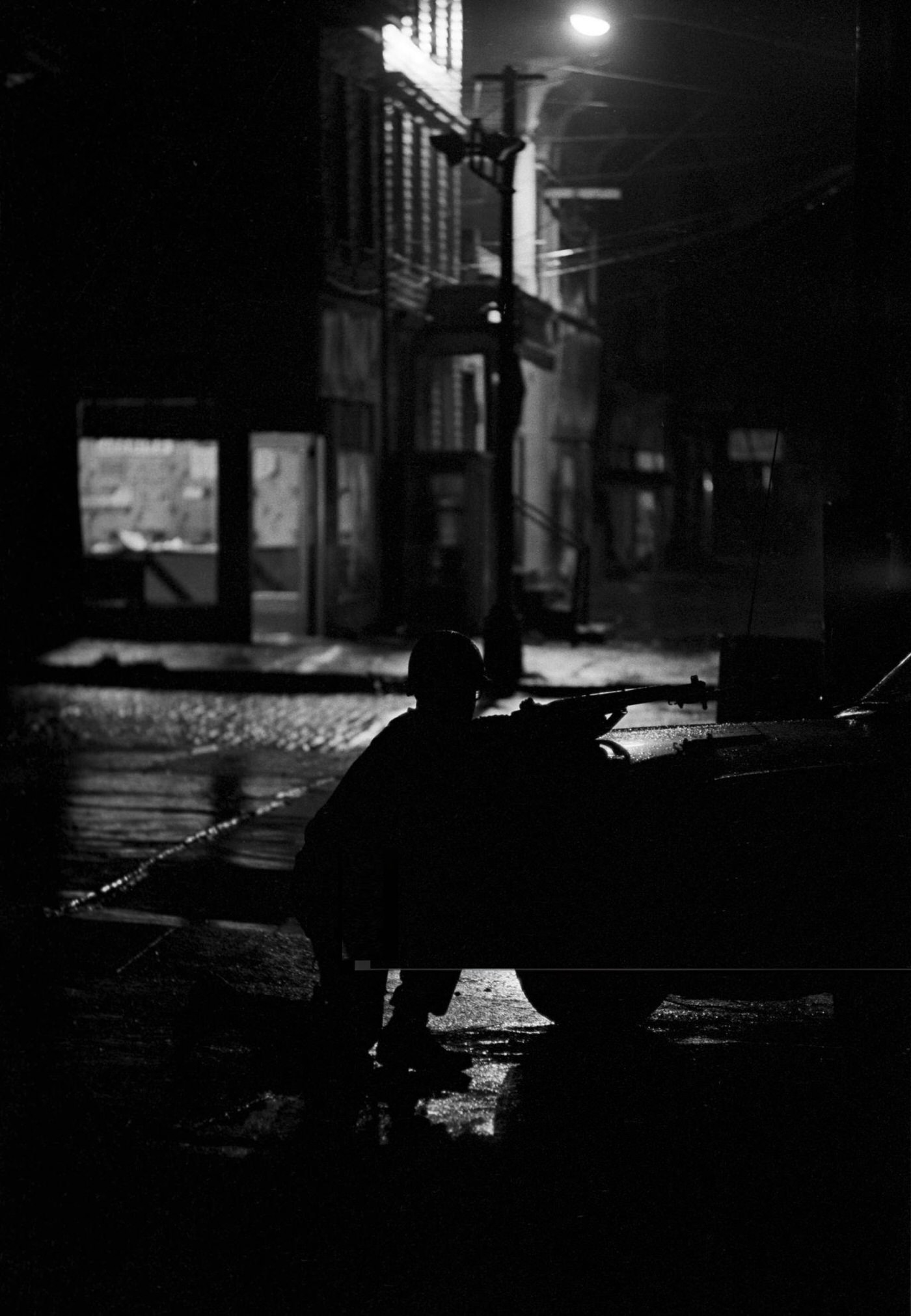 A National Guard soldier crouched behind a car, gun pointed towards the street at night