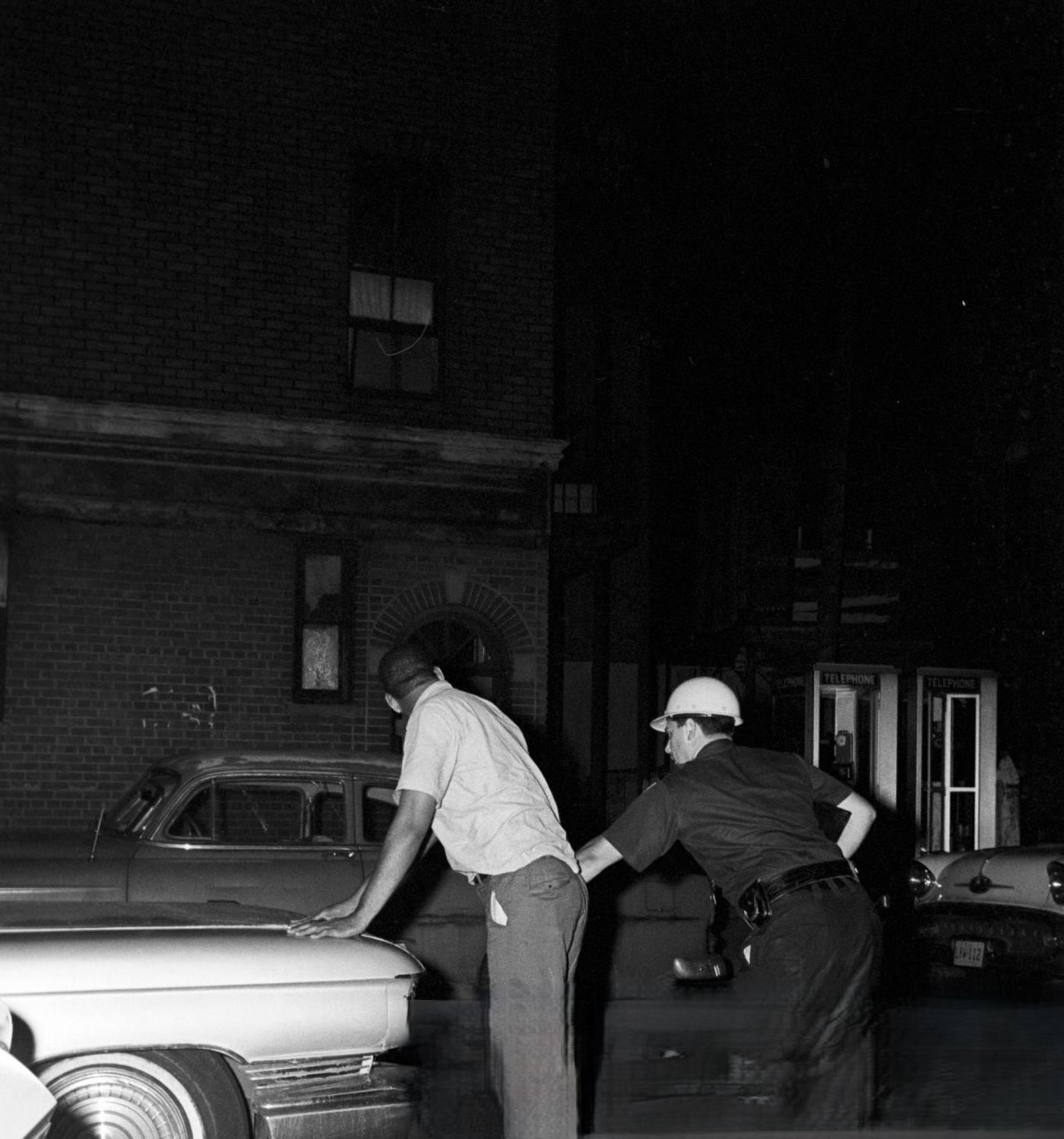 A National Police officer searches an African American man in front of a parked car