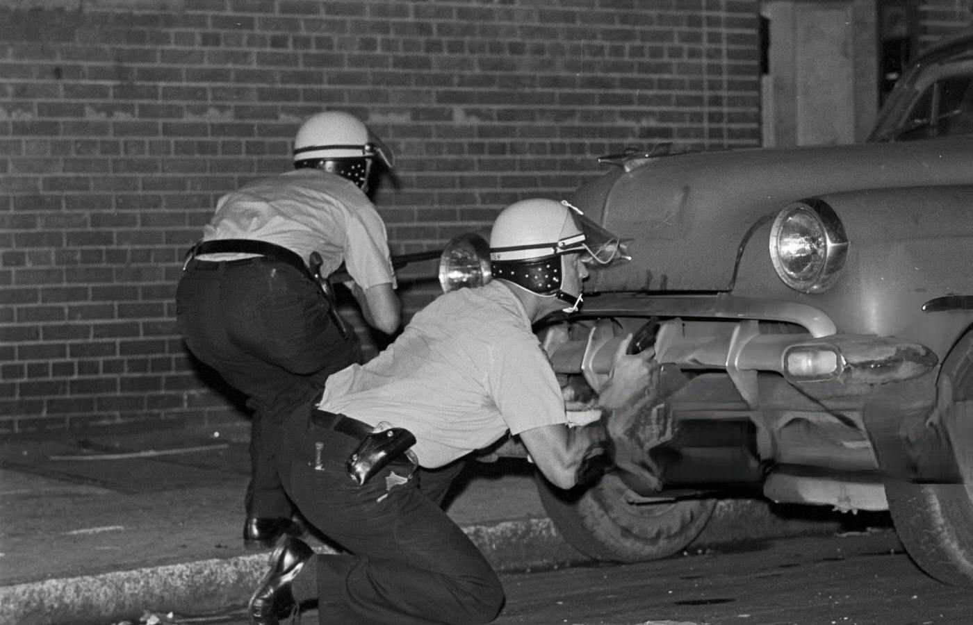 Two police officers in riot helmets take cover behind a parked car