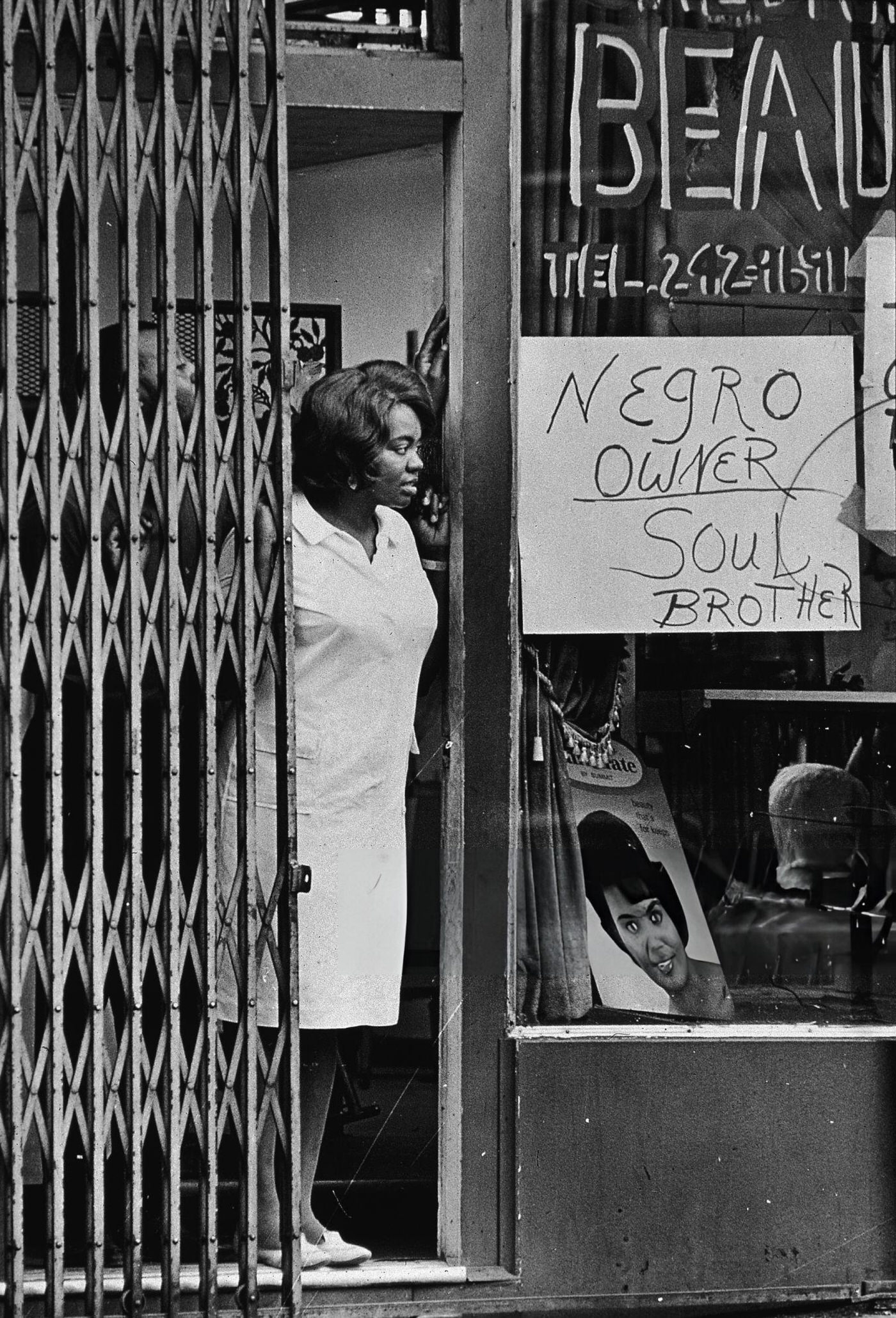 A Black woman peers from beside a gate in the door of a store with a sign in the window reading 'Negro owner,' 1967
