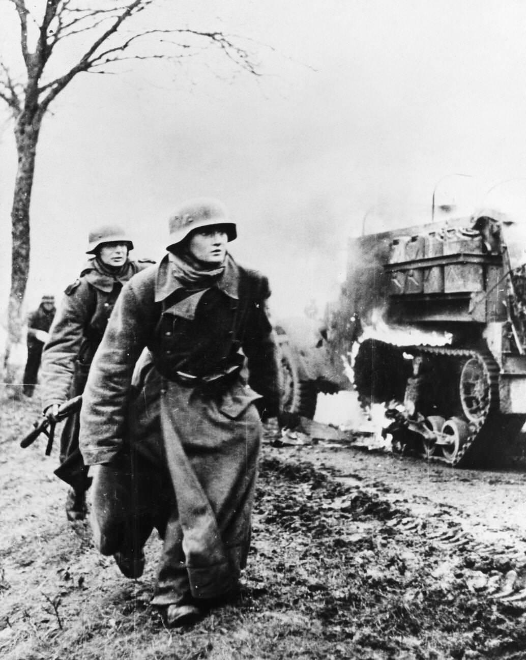 German troops passing by a burning US Army vehicle as the German infantry advanced during the Battle of the Bulge, possibly in Poteau, Belgium, 1944-1945.