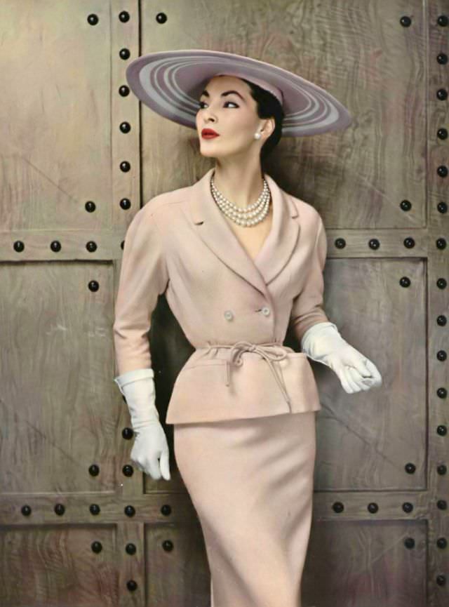 Lucie Daouphars in a lightweight glazed wool suit, the jacket has rounded shoulders and a shawl collar, tied at the waist with cord of the same material by Pierre Balmain, 1954.