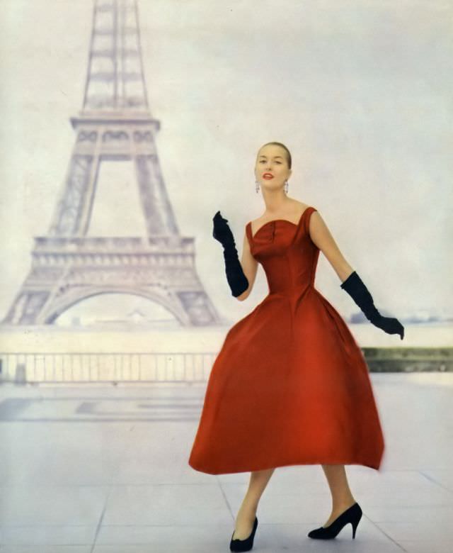 A model in a short little dance dress of red satin with an upturned circle neckline and belled skirt by Pierre Balmain, 1955.