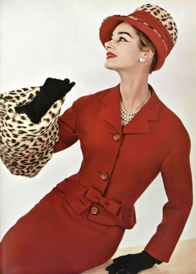 Jacky Mazel in a red Shetland wool suit, the fitted jacket has a small bow at the waist, worn with a leopard fur muff and hat by Pierre Balmain, 1956.