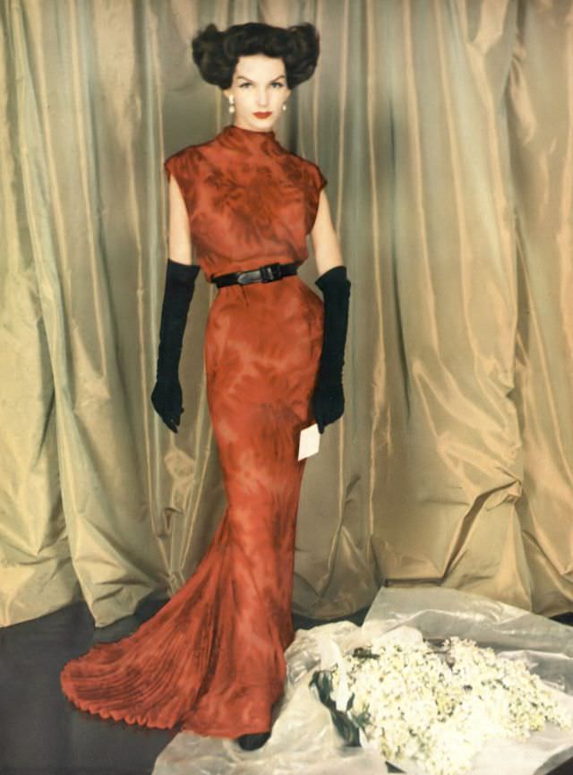 Joanna McCormick in a garnet red print chiffon sheath, bloused over a leather belt, an air-vent in the back and a fan pleat train, by Balmain, 1957.