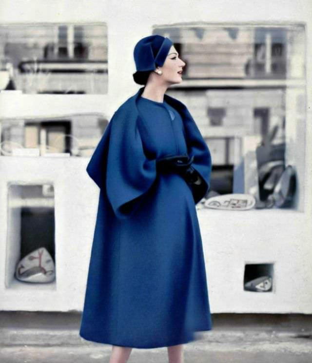 Simone d'Aillencourt in a roomy blue coat whose effect of a short cape at the back is given by the cut and sleeve setting, collarless and one large button-closure, by Pierre Balmain, 1957.