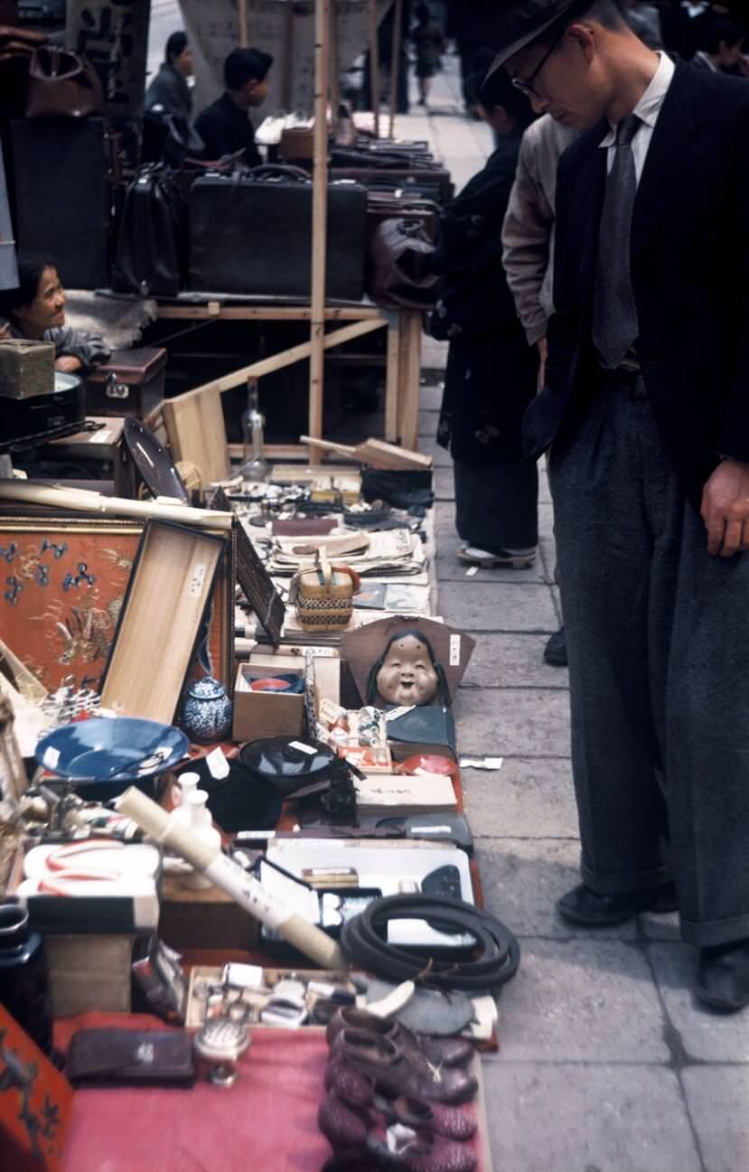 A Japanese man observing used merchandise for sale by a sidewalk vendor, 1950.