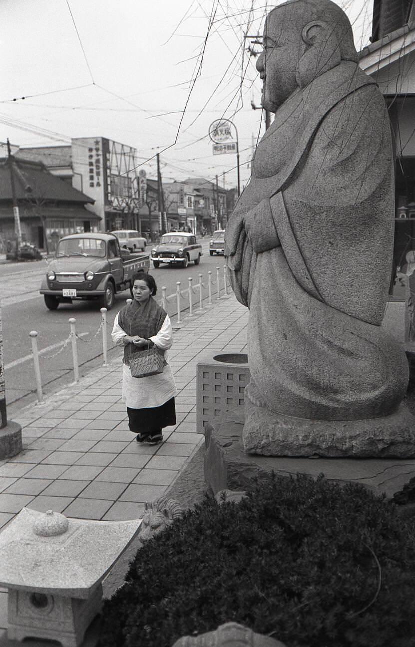 A housewife carrying a shopping bag walking in front of a statue of Ebisu at Tengenji, Tokyo, 1957.
