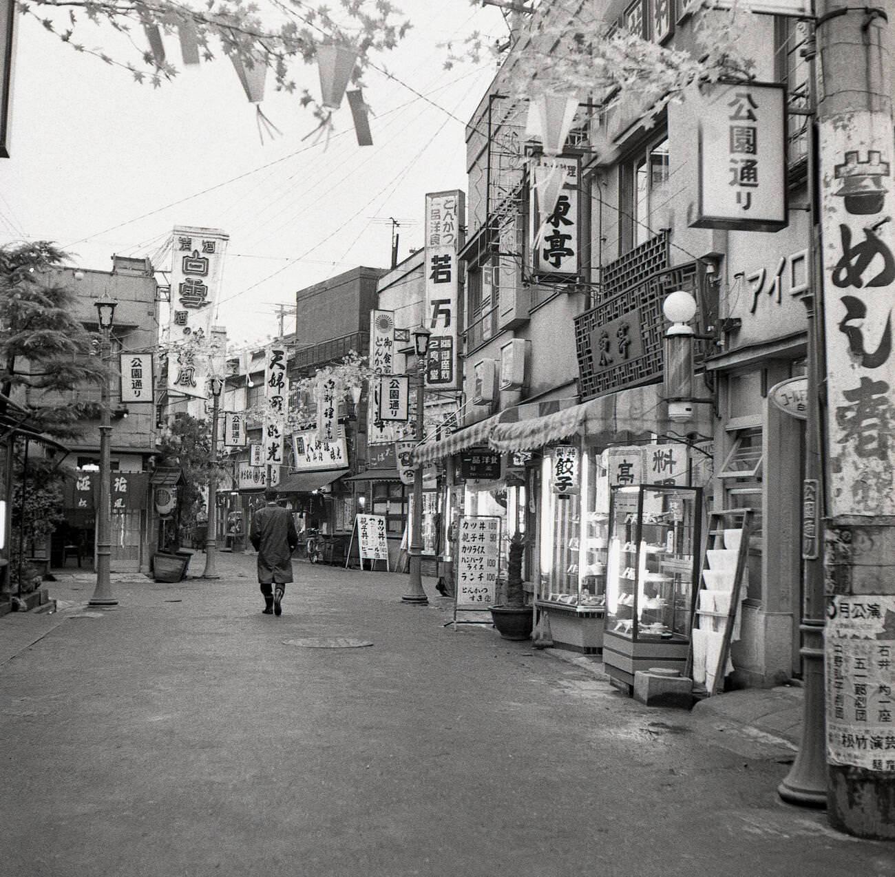 A man walking down pass the retail stores and buildings in a quiet side-street in Tokyo, 1950s.
