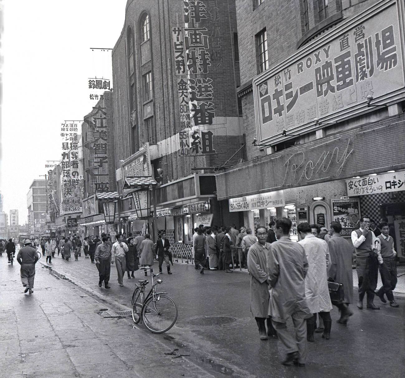 Men in raincoats walking along a wide pavement outside past the Roxy movie theatre, 1950s.