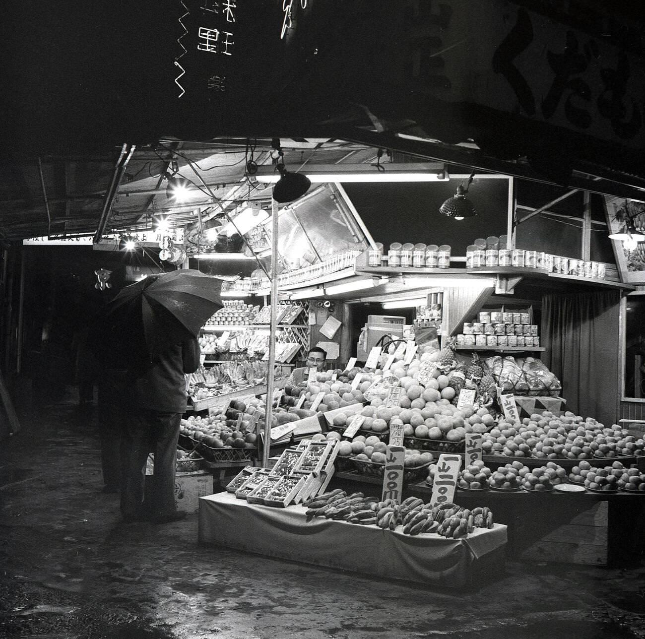 A wet evening and a man with an umbrella at an outdoor street stall selling fruit in Tokyo, 1950s.