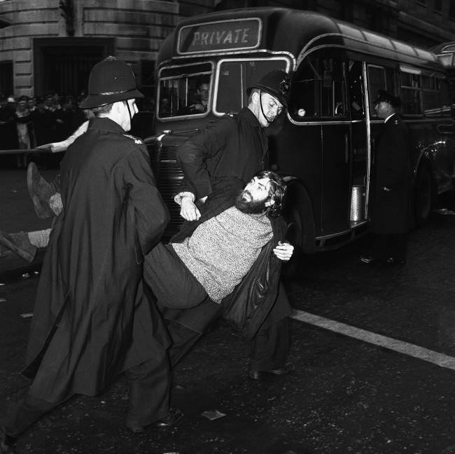 A long-haired young man, wearing a beard and beatnik style clothes, is carried to a waiting van by police after being arrested during the huge Ban-the-bomb rally in London's Trafalgar Square, United Kingdom, 1961.