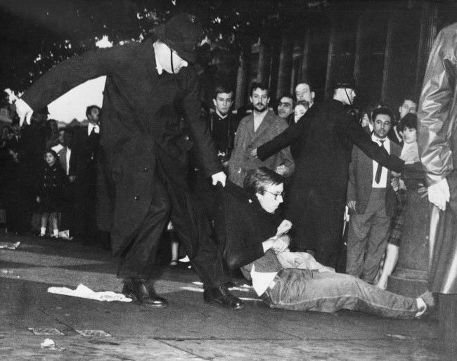 A young man in beatnik-style clothes is dragged along by a policeman, 1961.