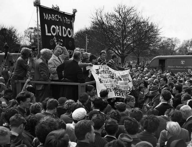 Two survivors of the Hiroshima atomic bomb hold up a banner as they join leaders of the Easter Ban-the-Bomb march on the dais in London's Hyde Park at the mass rally climaxing the 50-mile march from Aldermaston, 1962.