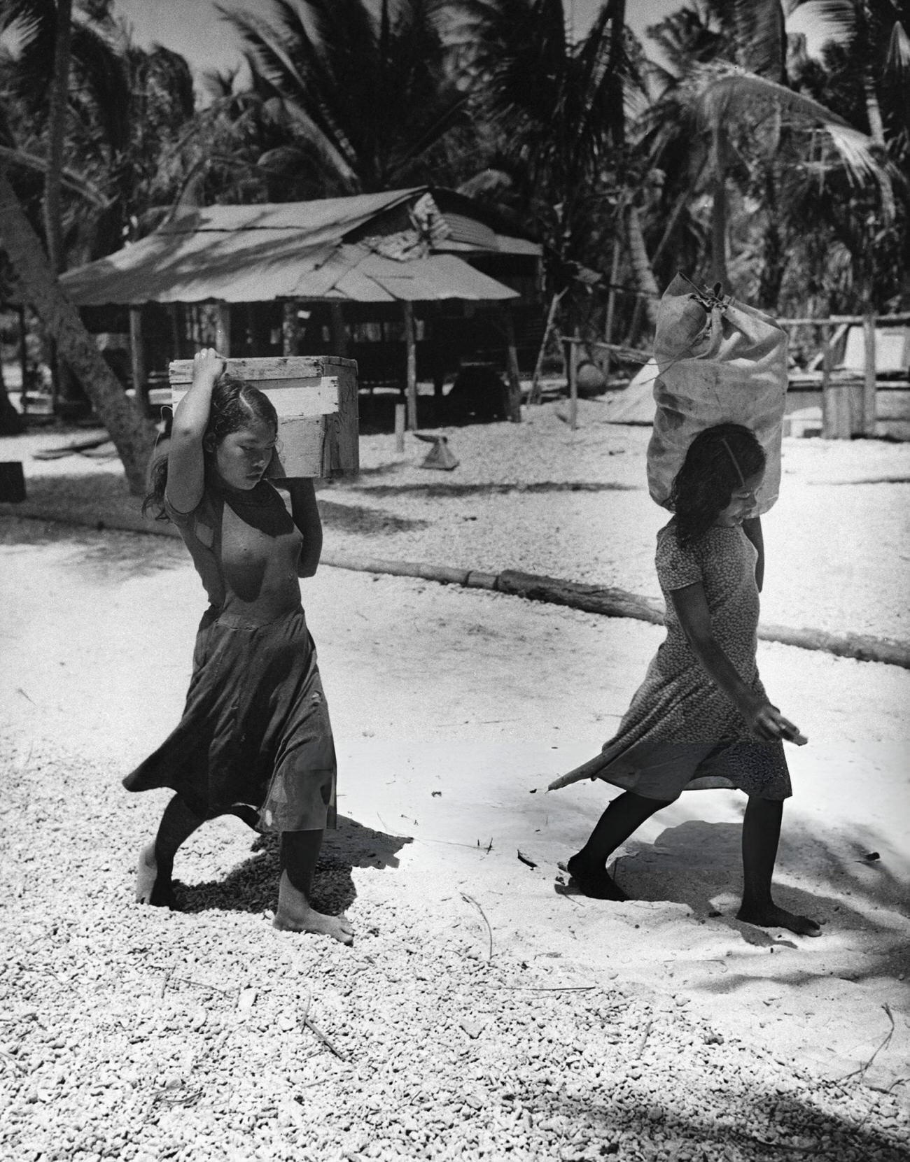 Two Bikini Atoll girls trudge away from their village carrying their possessions on their shoulders, 1946.