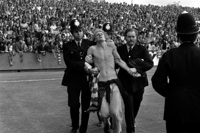 Stripped to the waist, a man is led away by police, 1970s.