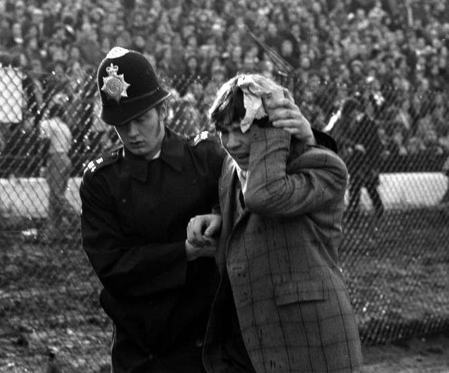 A Policeman escorts a young fan nursing an injured head, 1970s.