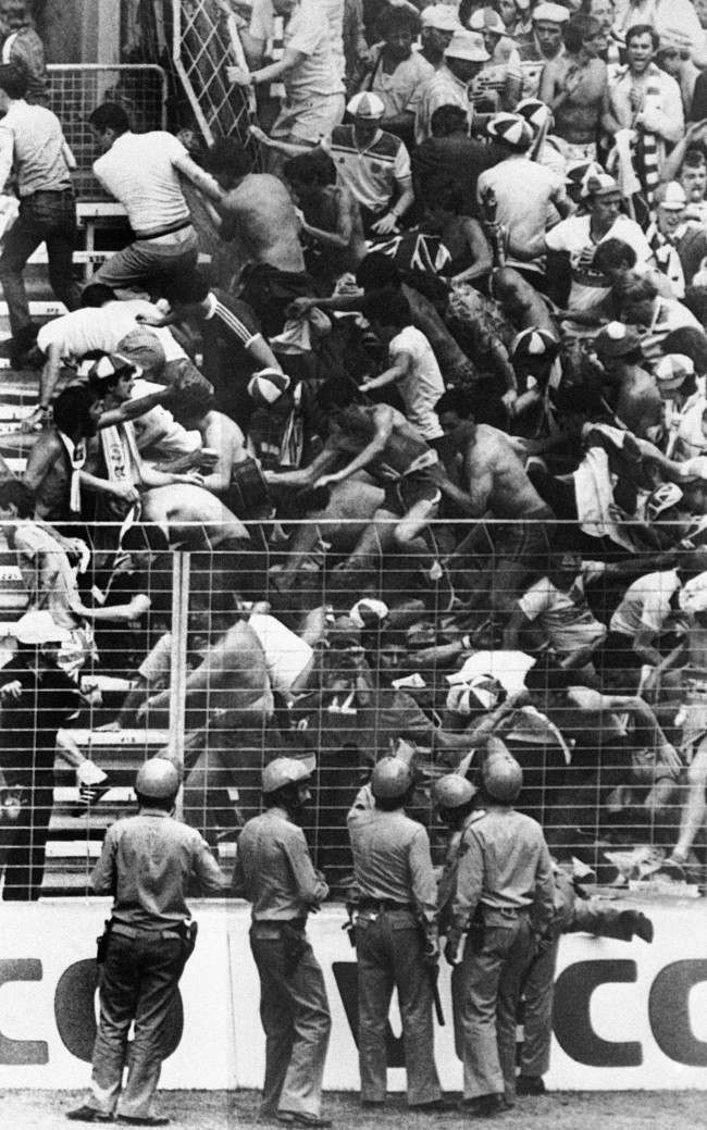 Spanish riot police with batons look on as England football fans tumble over barriers, 1980s.