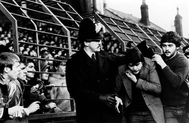 A Tottenham fan is escorted past the Anfield Road end by police, 1980s.