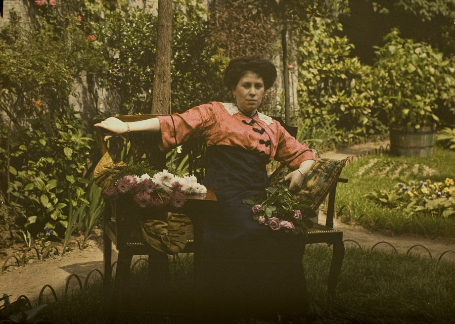 Mrs. Corbet on a garden bench with a flower bouquet, 1910