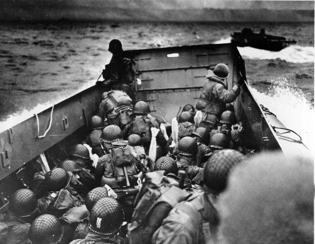 A U.S. Coast Guard landing barge, tightly packed with helmeted soldiers, approaches the shore at Normandy, France, 1944.