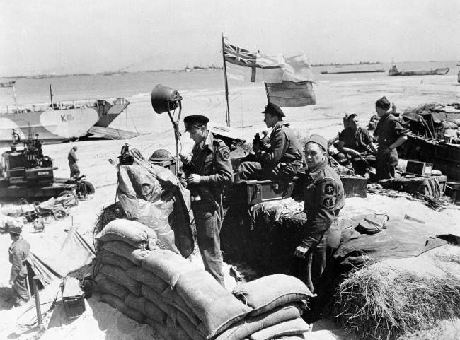 British troops make their way through low water and up the beach after leaving landing craft, 1944.