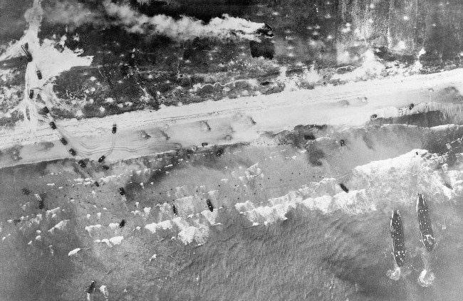 Allied troops come ashore in the surf and vehicles start inland on the German-held beach of Normandy, France, 1944.