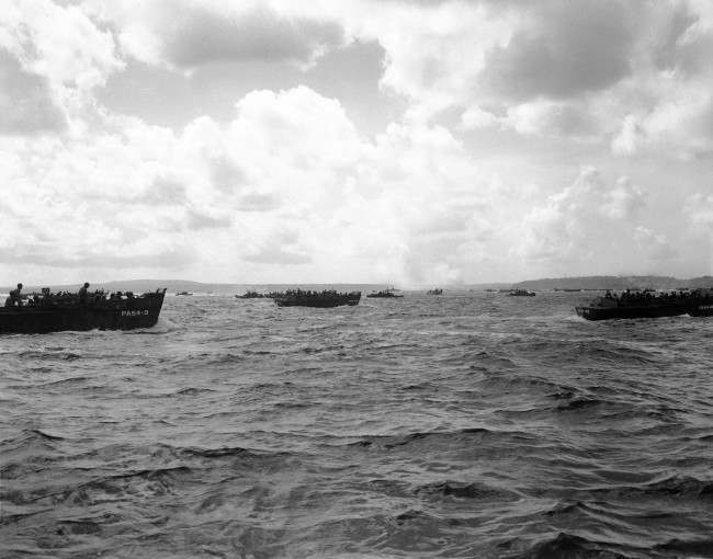 Smoke from Agana torpedo bomber targets are seen in the distance as landing craft race toward the Asan shore at Guam, 1944.