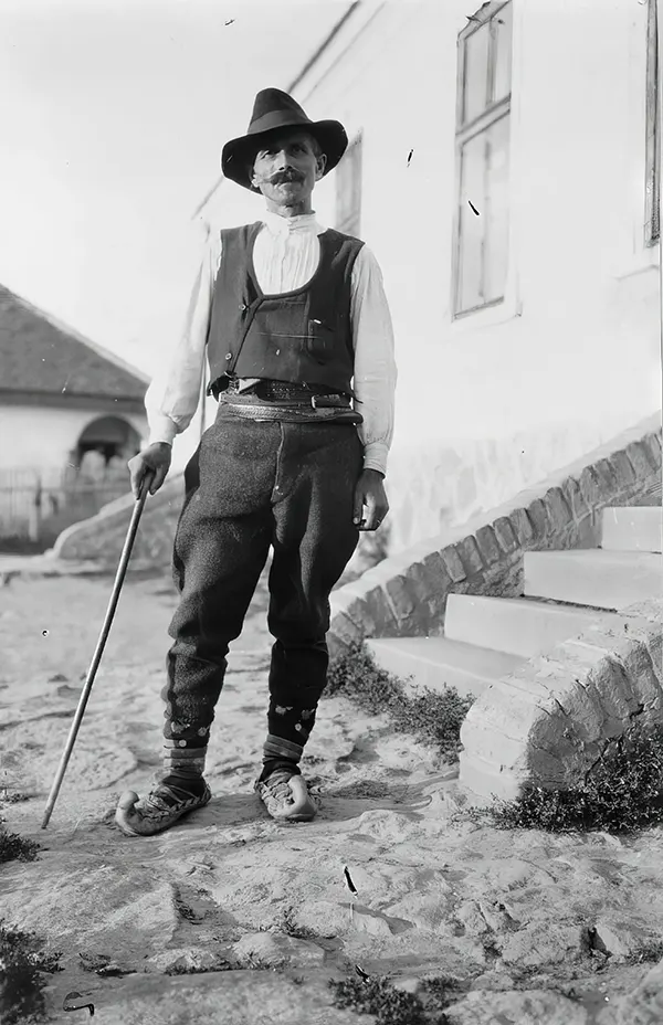 Serbian man wearing hat, vest, belted pants, and pointed toed shoes with cane standing outside of building in Yugoslavia, 1880-1924.