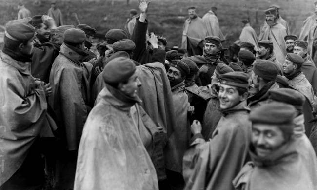 Cheerful German prisoners in forage caps and waterproof capes surround a member of a visiting News Agency in an effort to cadge cigarettes. Western Front, 1918.